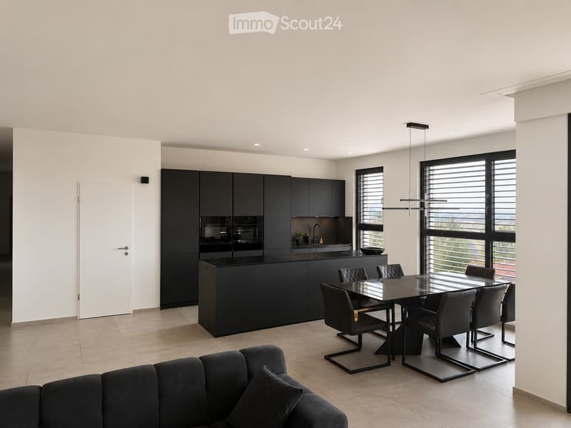 Modern, open-plan living and dining area with a black sofa, a glass dining table with six chairs, and a sleek black kitchen featuring cabinets, an oven, and a faucet. Large windows offer a view of greenery.