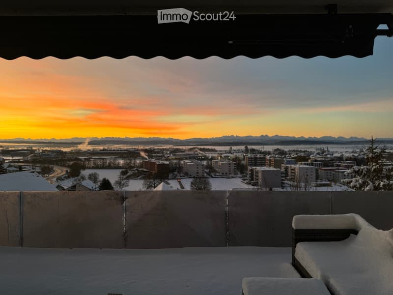 A snow-covered balcony with a view of a cityscape at sunset, featuring distant mountains and a frozen landscape.