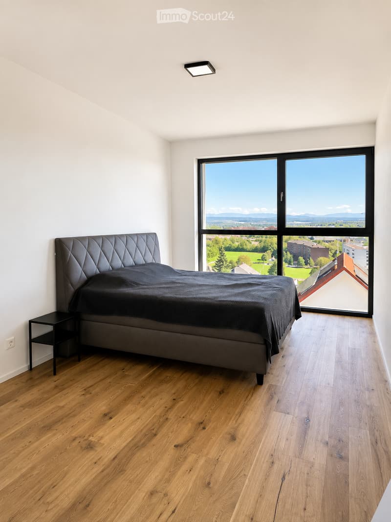 Modern bedroom with wooden flooring, large window offering a scenic view, a dark grey upholstered bed, and a small black nightstand.