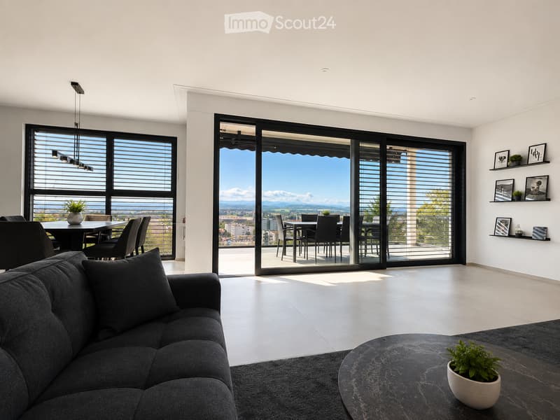 Modern living room with large windows and sliding glass doors leading to a balcony, featuring a grey sofa, coffee table, dining area, and decorative shelves with picture frames and plants.