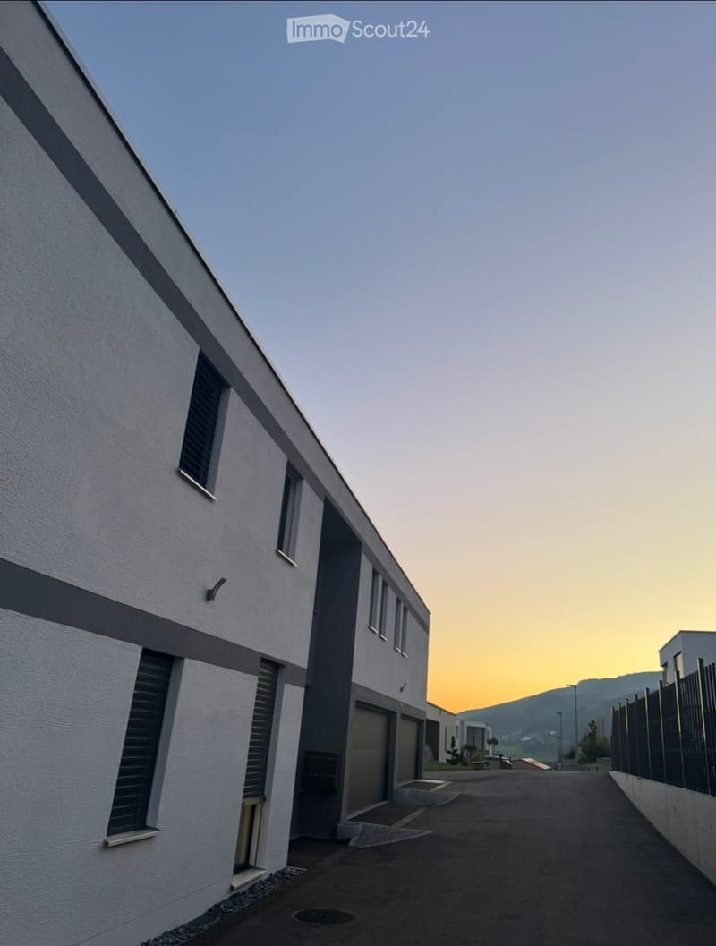 Modern two-story building with white and grey facade, multiple windows, and a garage entrance. The building is situated in a paved area with a fence and distant mountains visible in the background.
