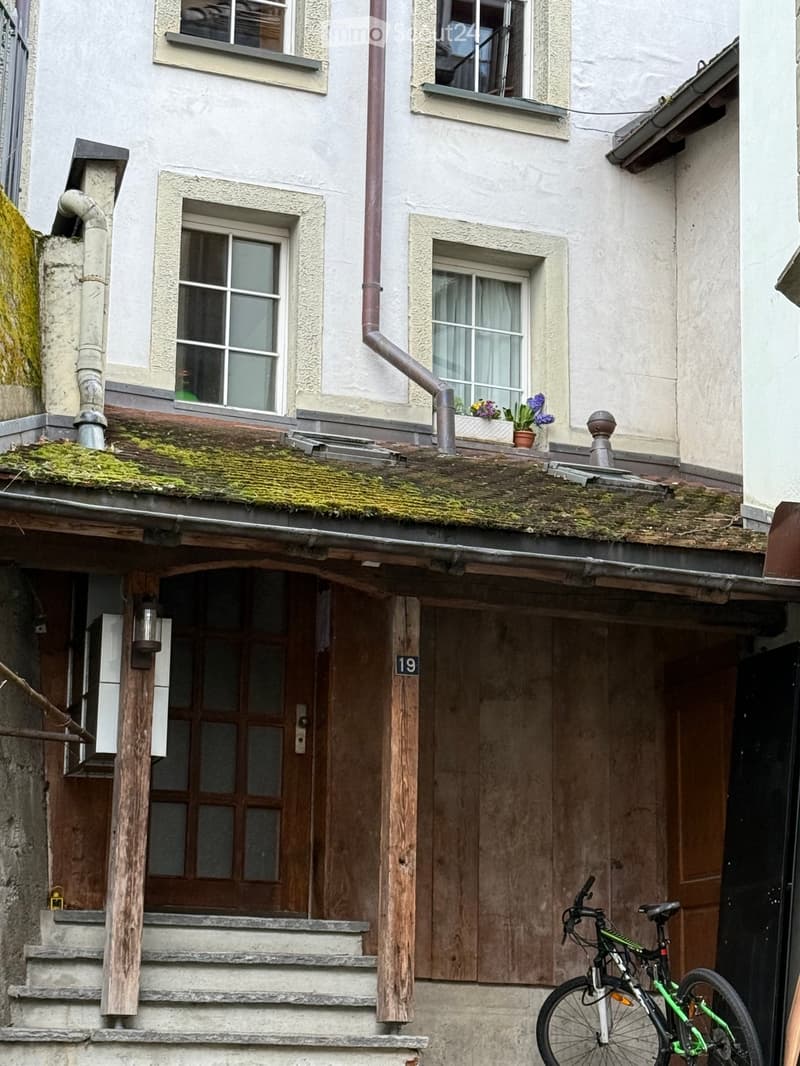 Two-story building with white facade, multiple windows, wooden shutters, and a small balcony with potted plants. A bicycle is parked near the entrance.