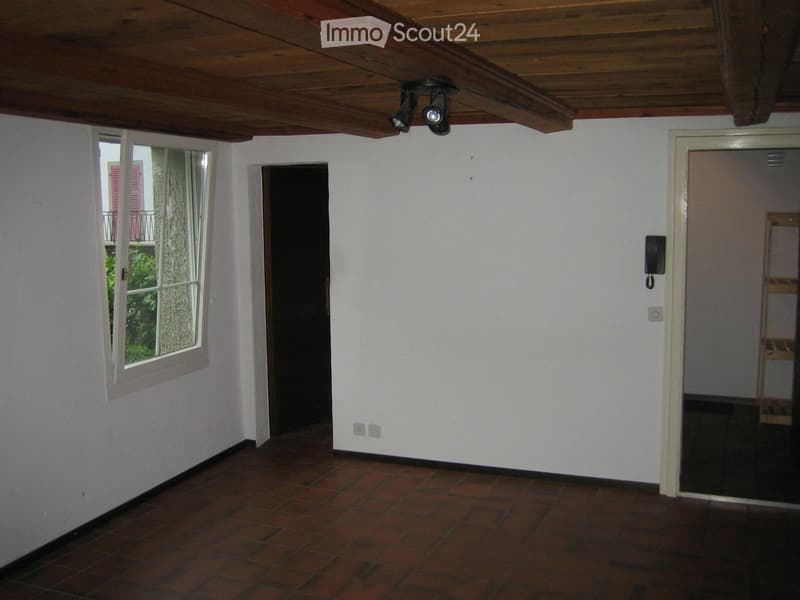 Empty room with exposed wooden ceiling, terracotta tiled floor, white walls, and a large window with shutters. There is a door leading to another room and a telephone mounted on the wall.