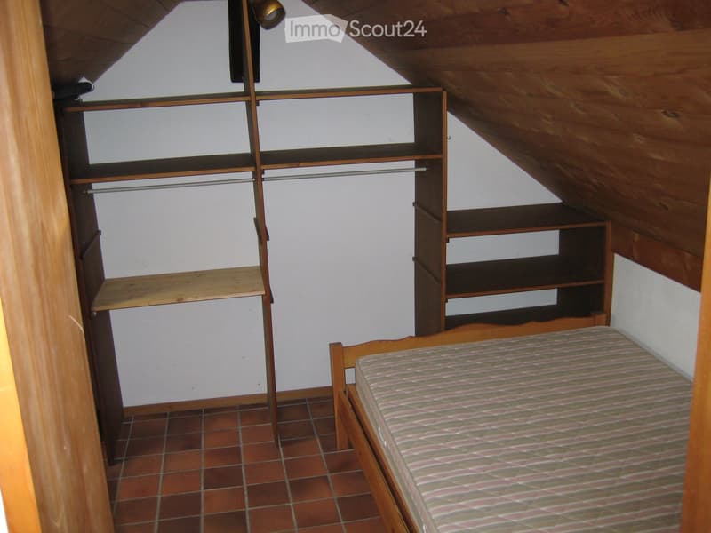 Bedroom with a single bed, wooden wardrobe, and built-in shelves. The room features exposed wooden beams on the ceiling and tiled flooring.