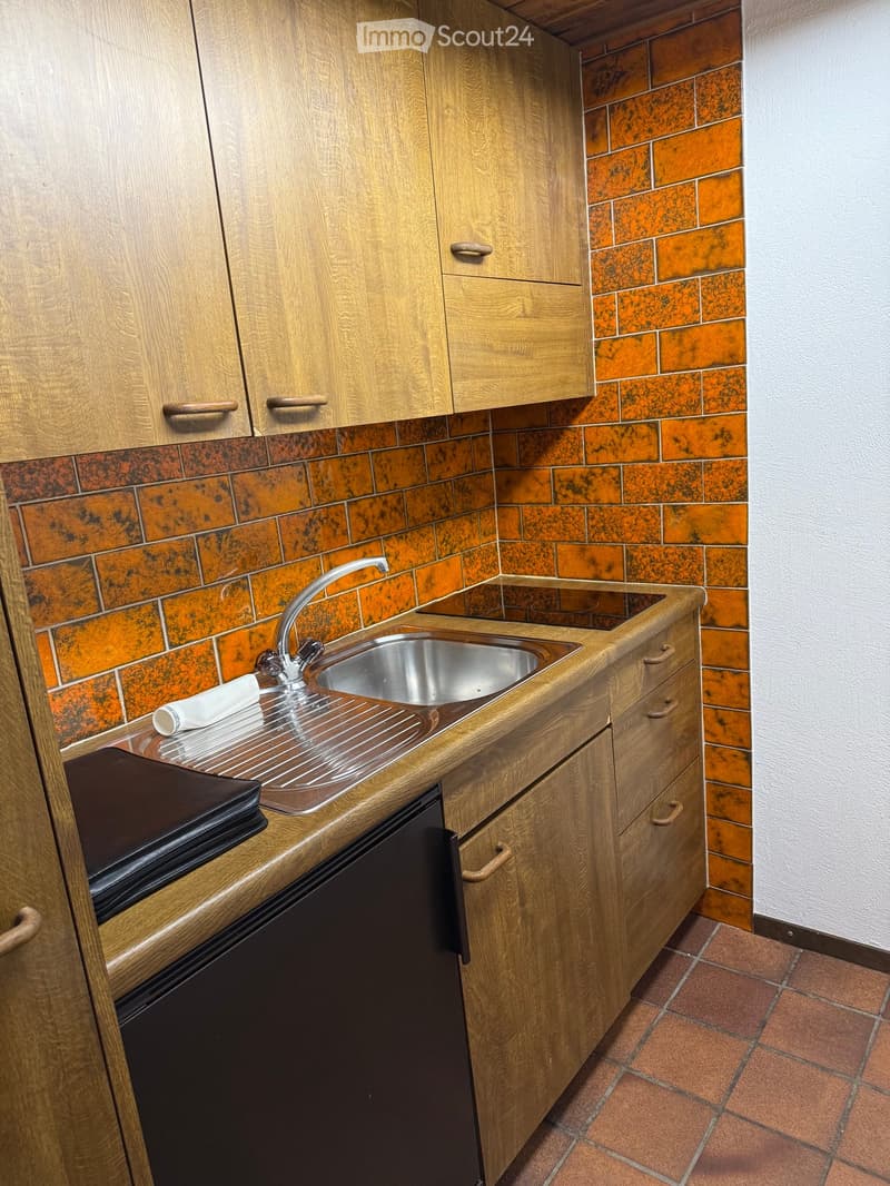 A compact kitchen with wooden cabinets, a stainless steel sink, and a refrigerator. The walls are tiled in a brick pattern, and the floor is tiled in a reddish-brown color.