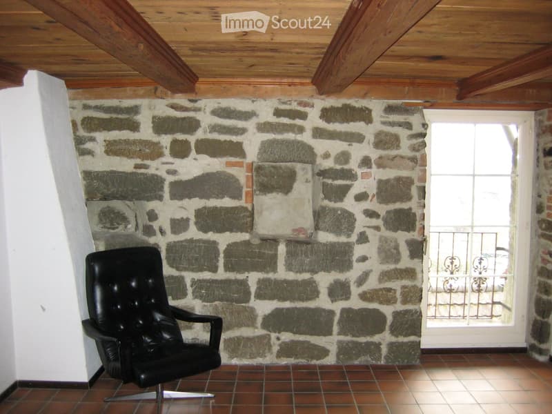A room with a stone wall, wooden ceiling, tiled floor, and a single black leather chair.