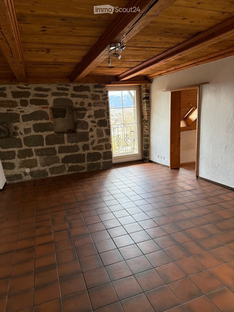 Empty room with exposed wooden beams on the ceiling, tiled floor, stone wall, and large window with a view.