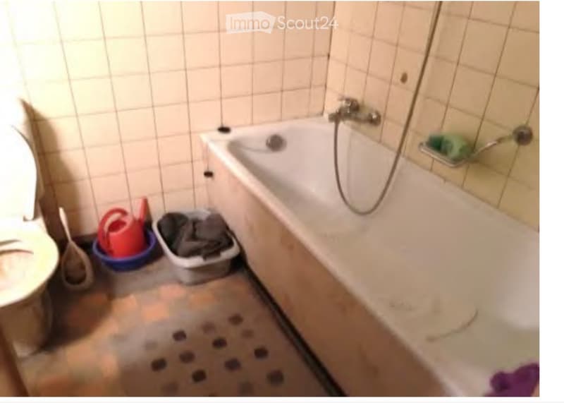 A small bathroom with a bathtub, toilet, and a shower hose attached to the wall. The floor is wet, and there are buckets and a trash bin near the bathtub.