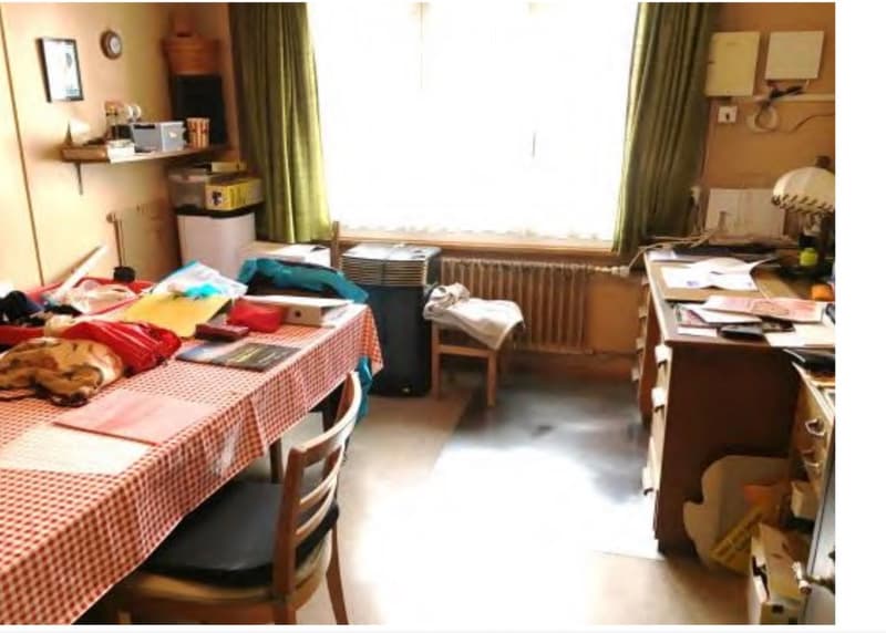A furnished room with a table covered in a checkered tablecloth, a chair, a desk with papers and a lamp, shelves with various items, and a radiator under a window with green curtains.