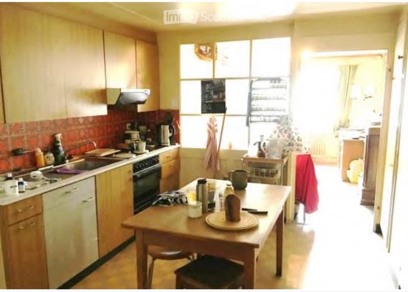 The image shows a well-lit kitchen with wooden cabinets, a central island table, and various appliances including a sink, oven, and dishwasher. The kitchen features a tiled backsplash and a window that allows natural light to enter. There is a small dinin