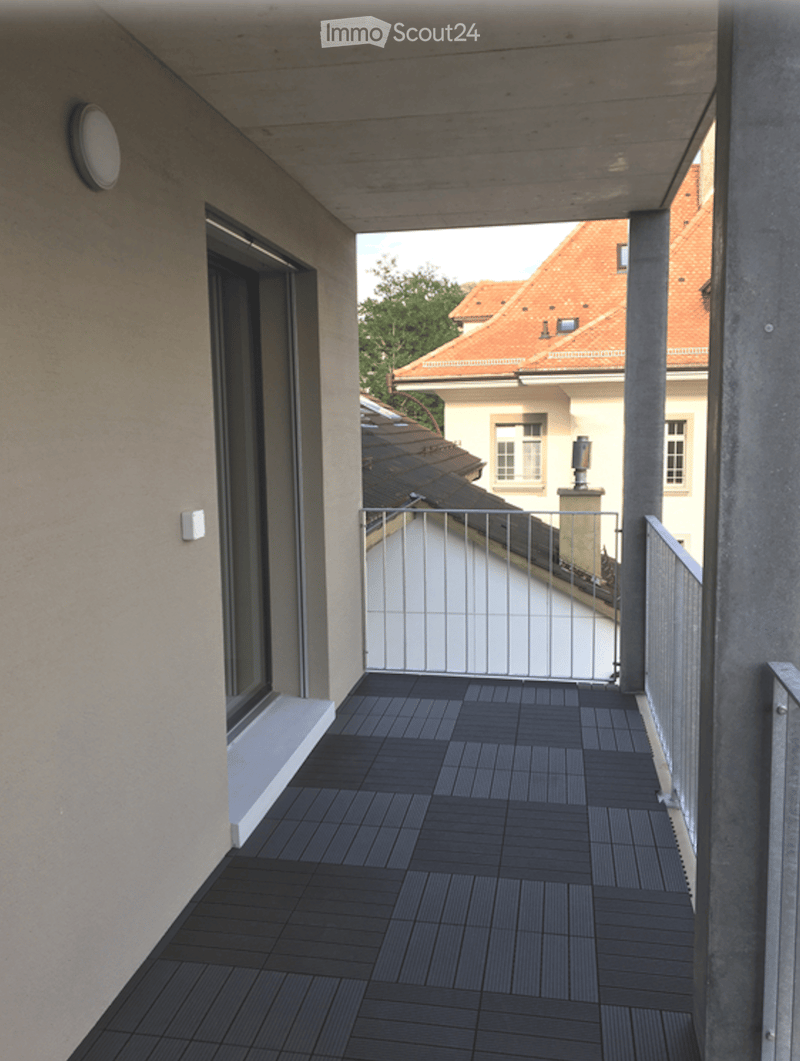 Modern balcony with dark wooden decking, metal railing, and a view of neighboring buildings with red-tiled roofs.