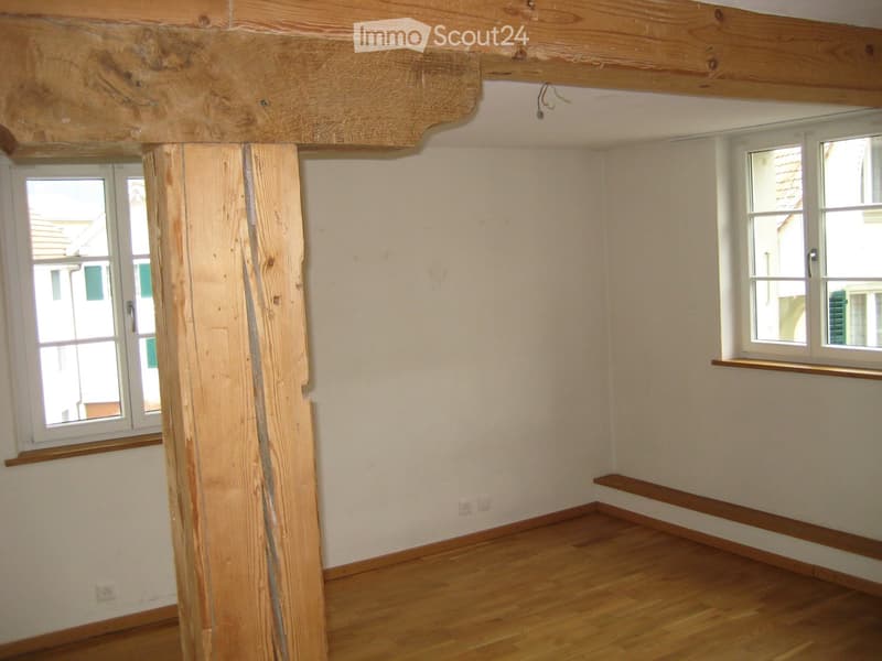 Empty room with wooden floor, exposed wooden beam, and windows with wooden sills.