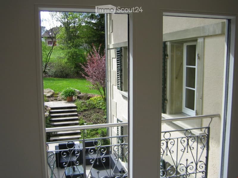 Balcony overlooking a well-maintained garden with steps leading down, featuring a metal railing and a view of the house exterior.
