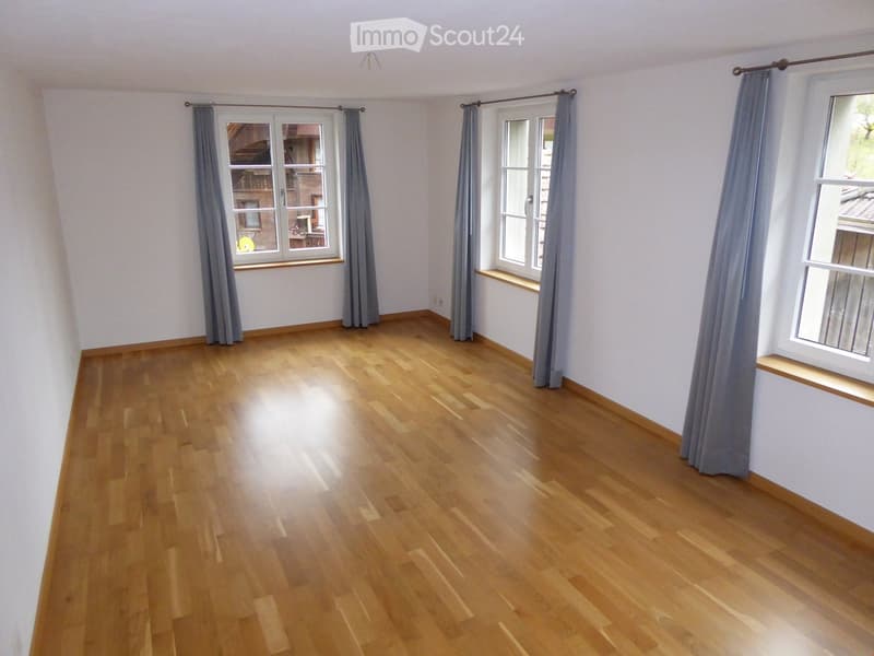 Empty room with wooden flooring, white walls, and three windows with grey curtains. The space is completely unfurnished.