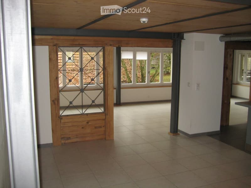 Empty room with tiled floor, white walls, wooden ceiling beams, and large windows with metal-framed glass panels.