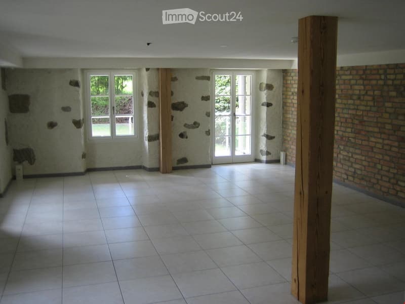 Empty room with tiled floor, exposed wooden beams, and large windows and doors leading to an outdoor area.