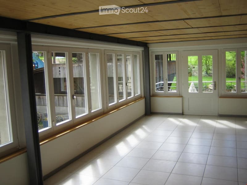 Bright, enclosed balcony with large glass windows and doors, white tiled floor, wooden trim, and a view of a green garden.