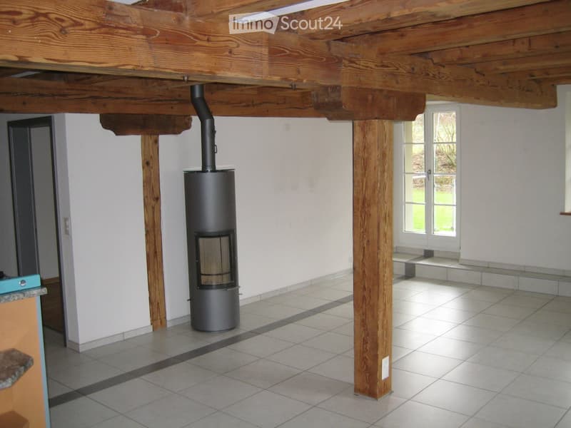 Empty room with exposed wooden beams, tiled floor, and a wood-burning stove. Natural light from a window on the right side.