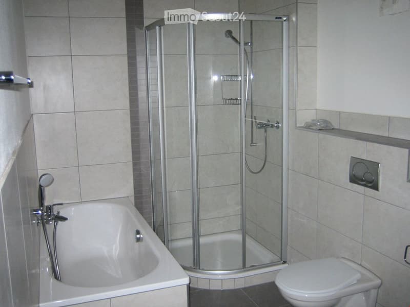 Modern bathroom with a bathtub, toilet, and a glass-enclosed shower stall. The walls are tiled in light beige, and the floor is dark.