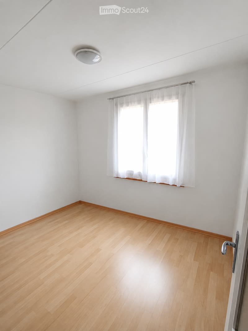 Empty room with light wooden flooring, white walls, a window with white curtains, and a ceiling light fixture.