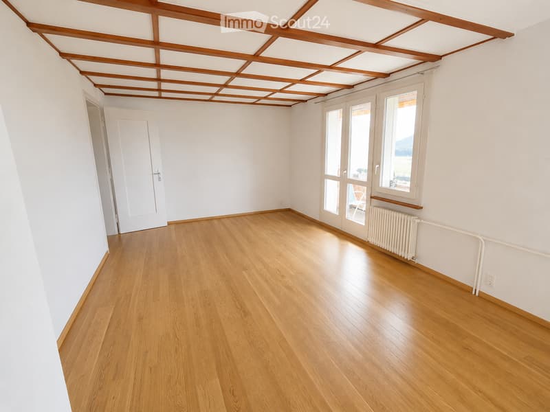 Empty room with wooden flooring, white walls, and large windows with white frames. The ceiling features exposed wooden beams.
