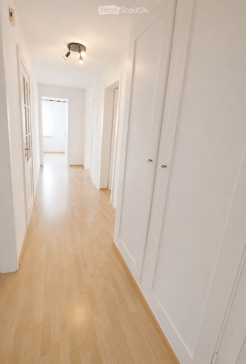Empty hallway with light wooden flooring, white walls, and several closed doors. A ceiling light fixture is visible.