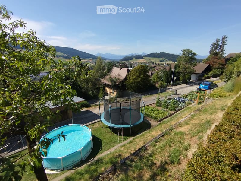 Aerial view of a garden area with a small swimming pool and a trampoline, surrounded by greenery and trees, with a scenic mountain backdrop.