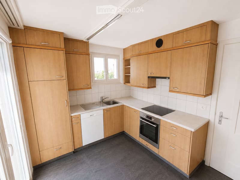 Modern kitchen with light wooden cabinets, white countertops, stainless steel sink, oven, and dishwasher. Features a window and built-in storage.