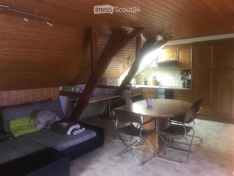 A cozy, loft-style living and dining area with exposed wooden beams, a sofa, a round dining table with chairs, and an open kitchen area with cabinets and appliances.