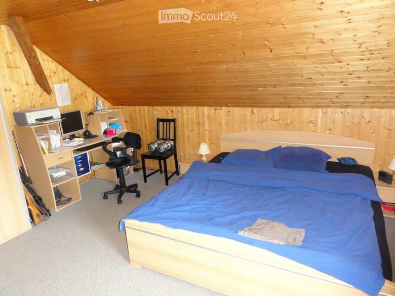 Cozy attic bedroom with wooden walls and ceiling, featuring a double bed with blue bedding, a desk with a computer, a chair, and a small table with a lamp.