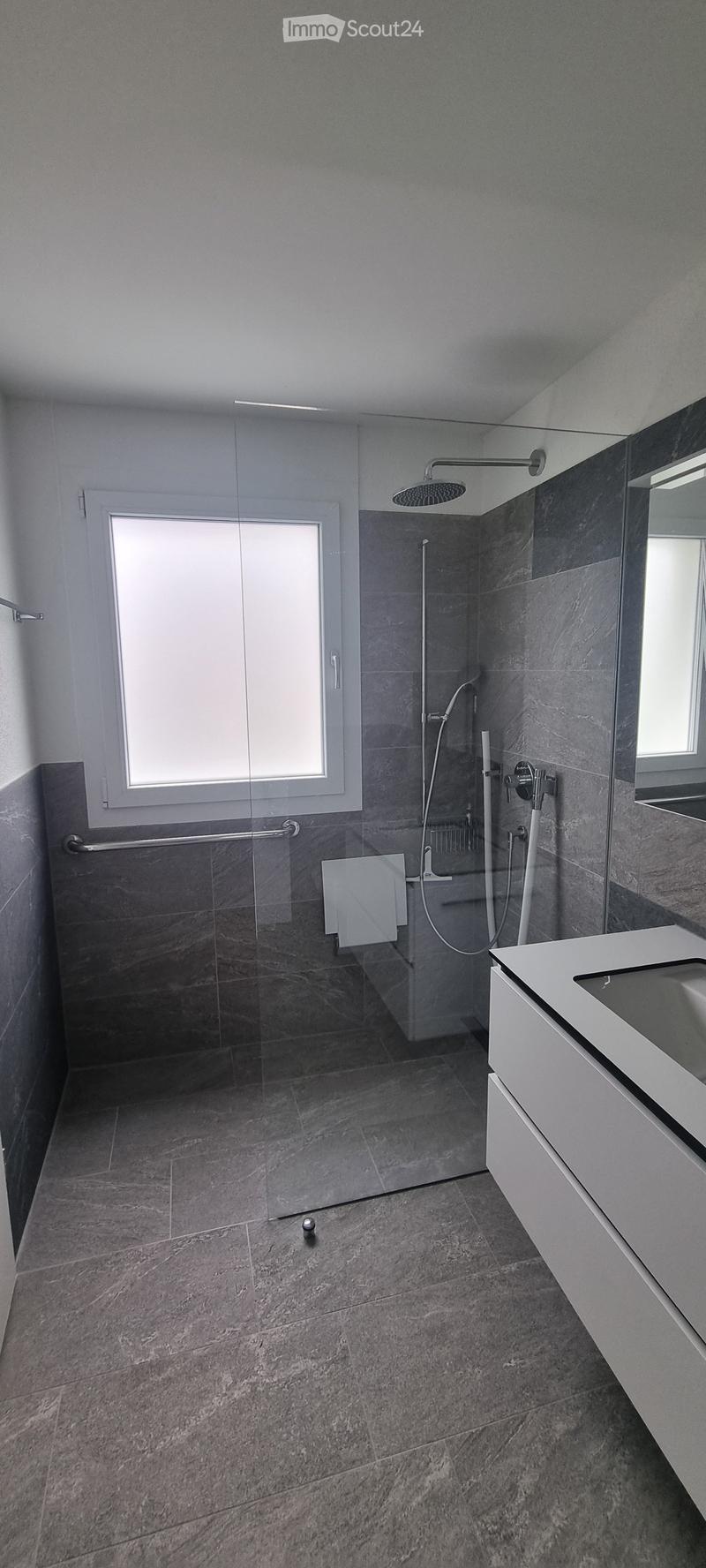 Modern bathroom with grey marble tiles, glass shower enclosure, white sink, and large window providing natural light.