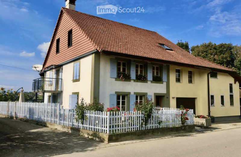 Two-story house with a red-tiled roof, light yellow walls, and blue shutters. Features a small balcony on the upper floor, a white picket fence, and a garden with flowers.