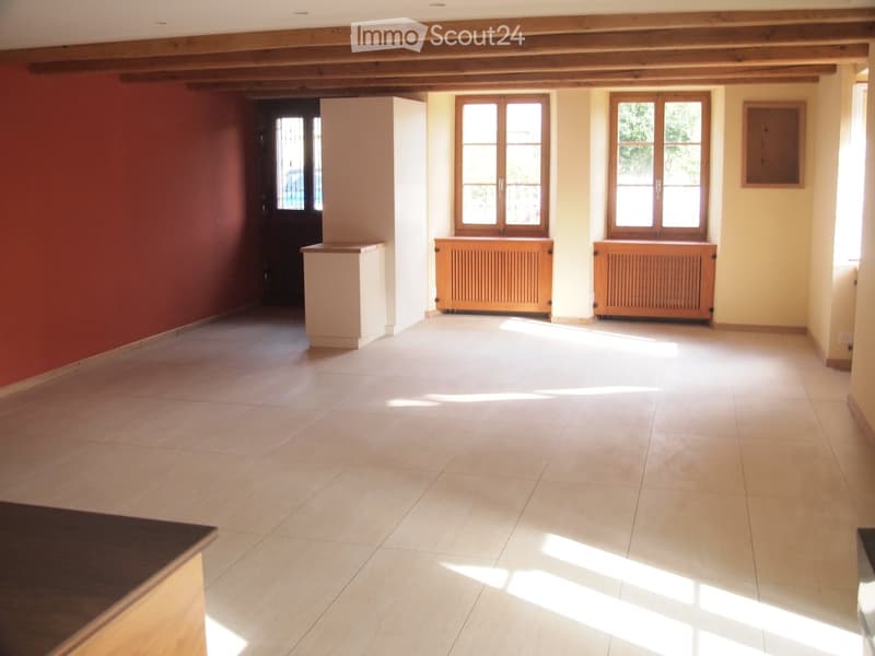 Empty room with tiled floor, wooden beams on the ceiling, and large windows allowing natural light to enter. There are radiators beneath the windows and a small partition in the corner.