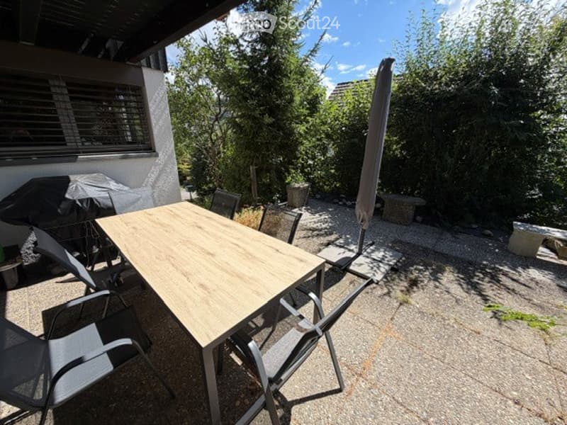 Outdoor patio area with a wooden table and several chairs, surrounded by greenery and trees. A large umbrella is partially open, and the area is paved with stone tiles.
