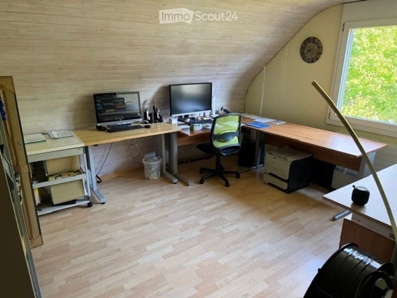Bright, wooden-floored room with a large desk, two computer monitors, a chair, and a printer. The room has large windows and a slanted ceiling typical of an attic space.
