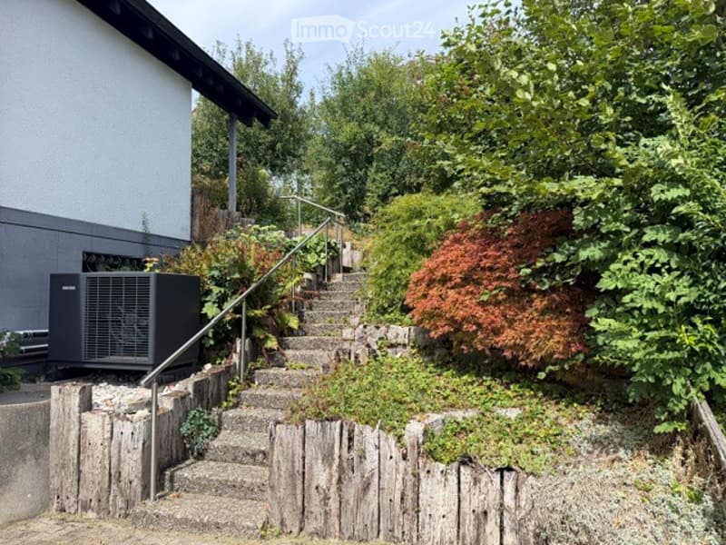 A stone staircase leads up to a house, bordered by a wooden fence and surrounded by lush greenery and trees.