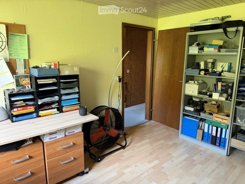 Bright room with light yellow walls and wooden flooring, featuring a desk with storage bins, a standing fan, and a tall shelving unit filled with books and office supplies.