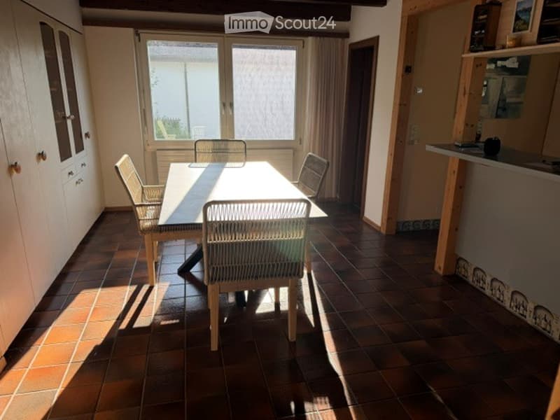 Bright dining area with a wooden dining table and four chairs, large windows with curtains, and a cabinet on the left side.