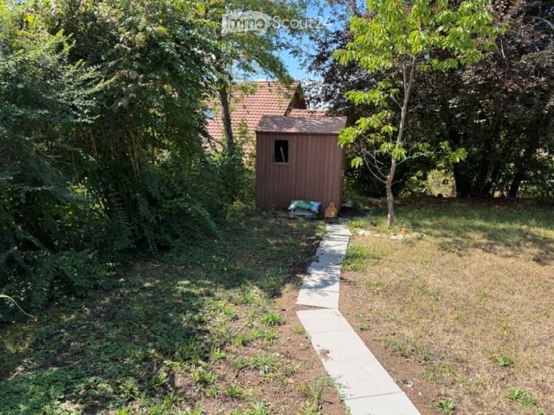 A small wooden shed is situated in a grassy yard surrounded by trees. A concrete pathway leads to the shed, and some bags and items are placed near the entrance.