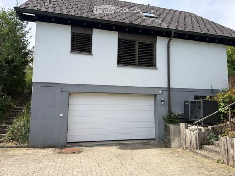 Two-story house with a gray garage door, white walls, and multiple windows. The roof is dark and sloped, and there is a small staircase leading up to the entrance.