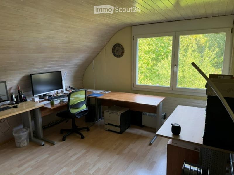 A well-lit attic office with wooden floors, large windows, and a slanted ceiling. The room features a desk with a computer monitor, chair, keyboard, mouse, and printer, along with a small table holding additional office supplies.