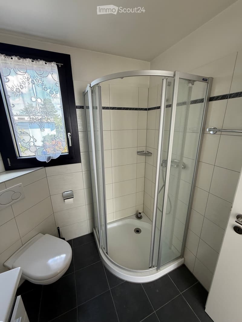 Modern bathroom with a corner shower stall, toilet, and window with a decorative curtain. The walls are tiled in light beige with black accents, and the floor is dark tiled.