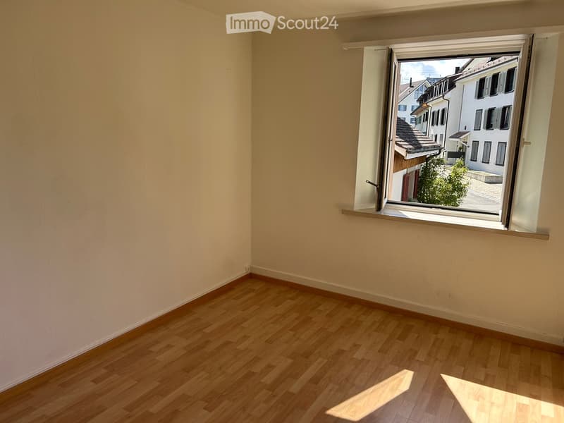 Empty room with wooden flooring and a large window showing a view of a town with buildings and a tree.