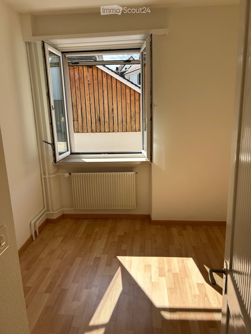 Empty room with wooden flooring, light-colored walls, a radiator, and a large window with a wooden blind partially visible outside.