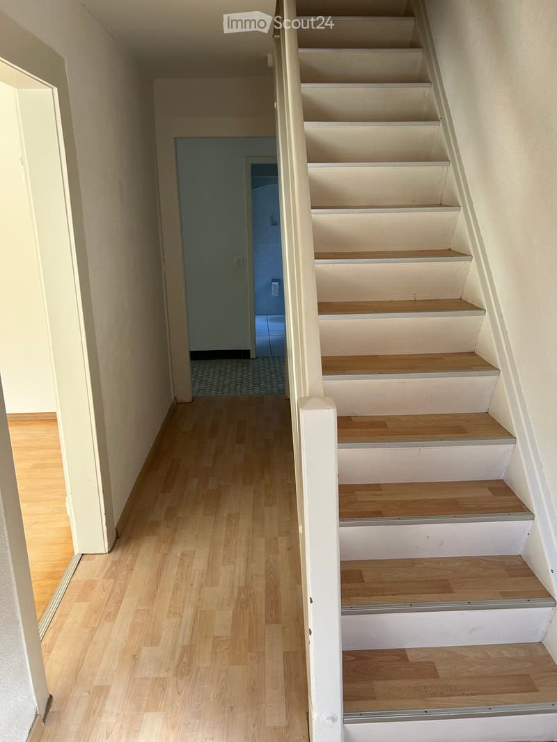 A narrow hallway with wooden flooring and a staircase leading upwards, featuring white walls and a door at the end.