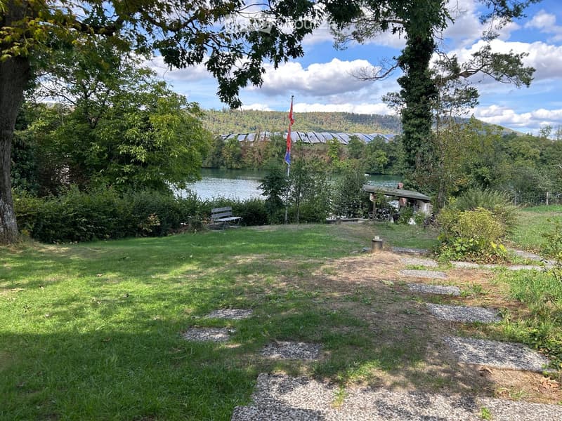 Lush green garden area with a bench, a small structure, and a view of a lake and surrounding trees.
