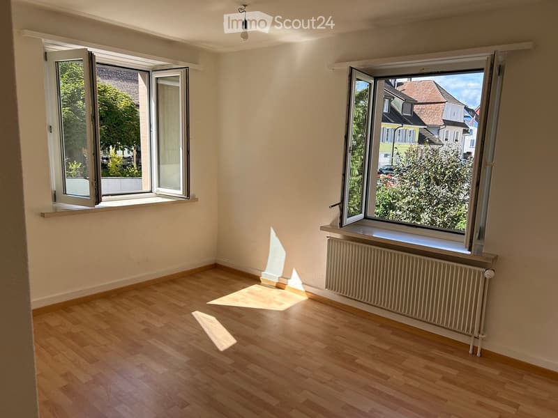 Empty room with wooden flooring, large windows, radiator, and natural light. No furniture or equipment present.