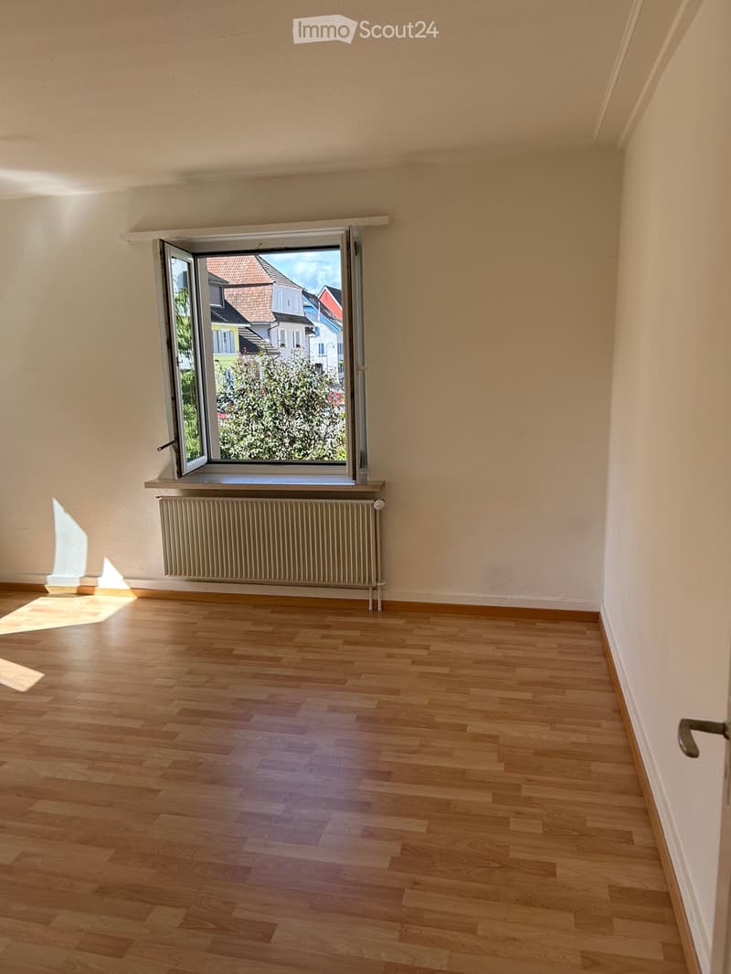 Empty room with wooden flooring, white walls, and a large window with a radiator below it. Sunlight streams in from the window, casting shadows on the floor.