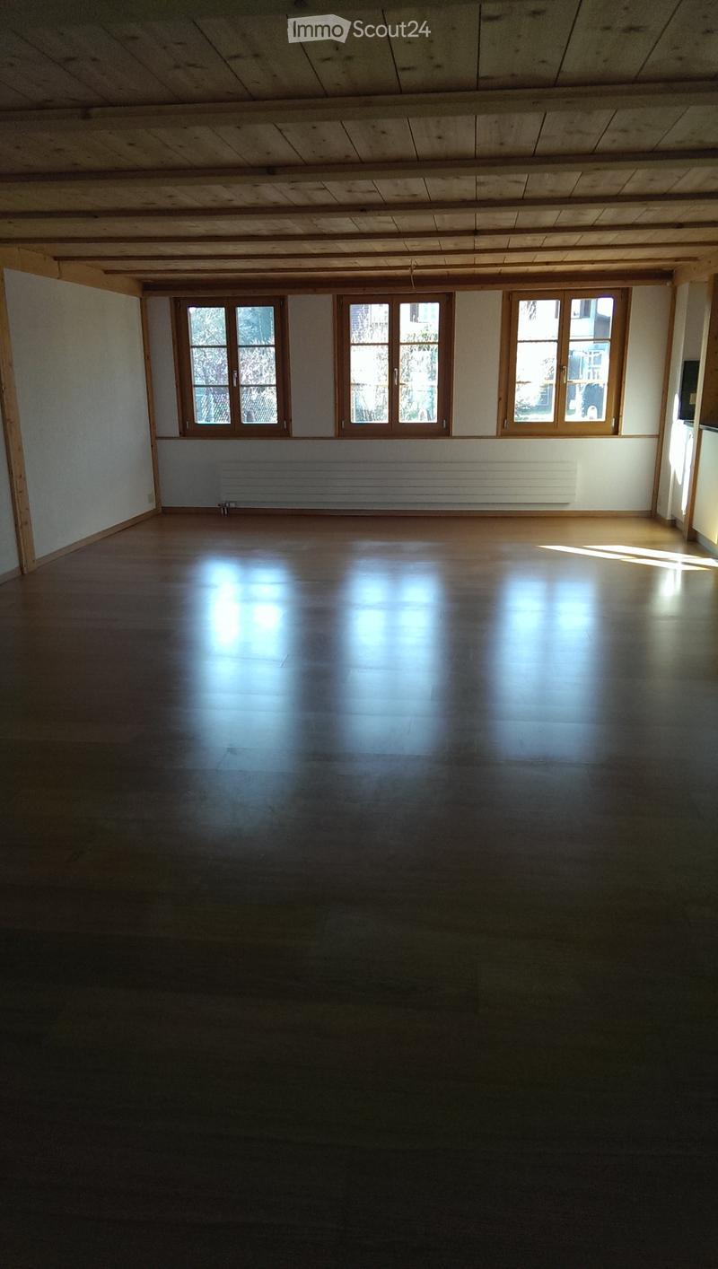 Empty room with wooden floor, white walls, large windows, and a radiator under the windows.