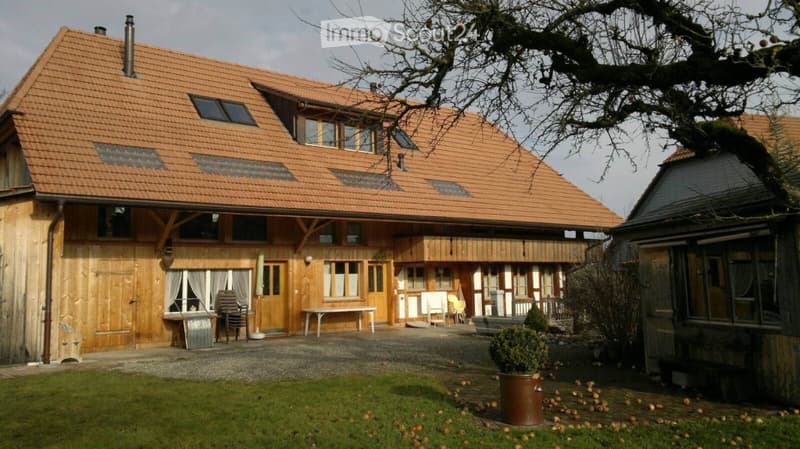 Two-story wooden house with a red-tiled roof, multiple windows, and a covered porch area with outdoor furniture.